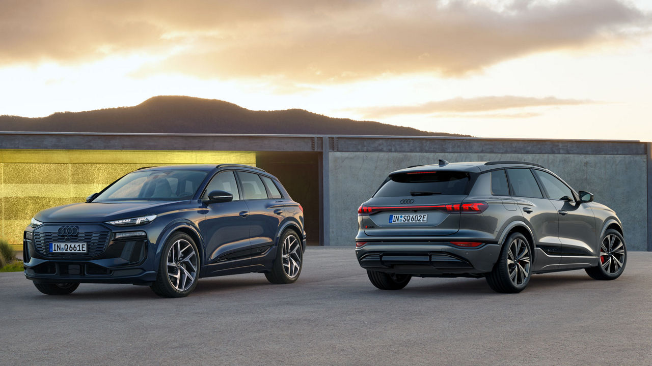 Two Audi Q6 e-tron parked in front of a yellow-grey wall against a mountain backdrop.